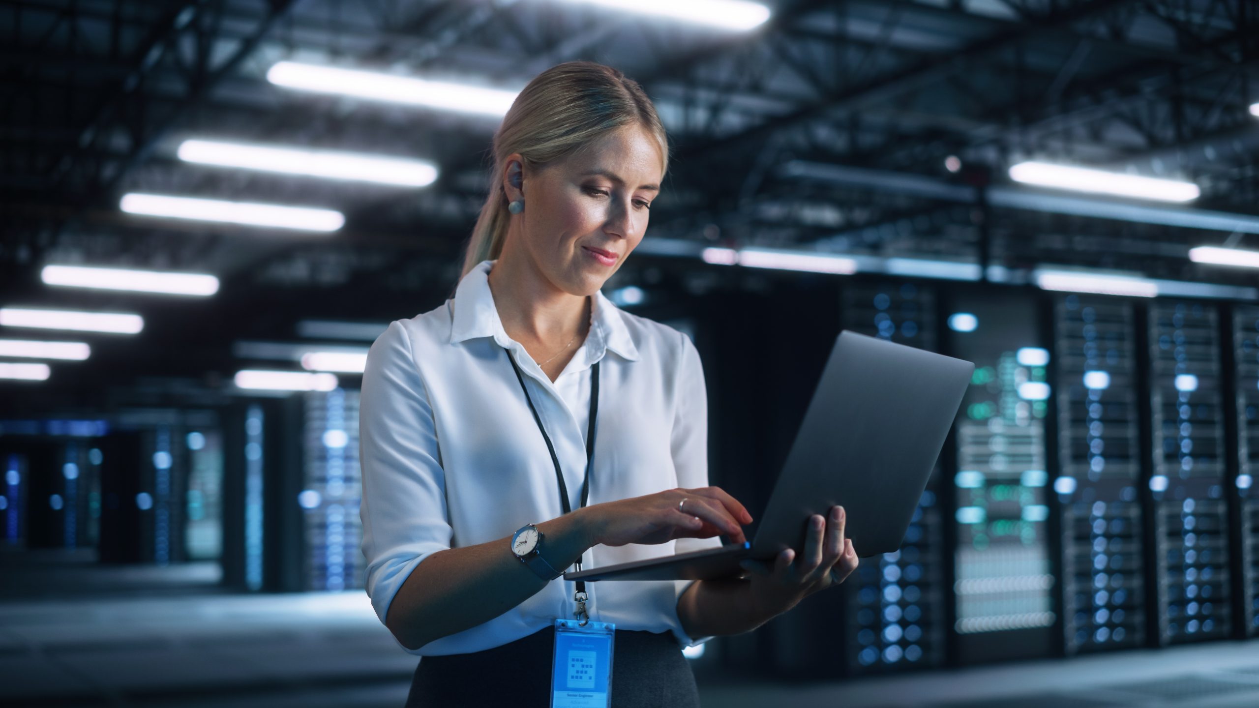 A blonde lady holding a laptop in front of a data center providing IT Management Support.